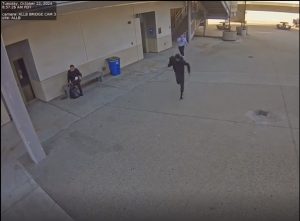 A security camera still image shows an outdoor area near a building with a person dressed in black running towards the camera. Another individual in a white shirt appears to be following them from behind. A third person is sitting on a bench against the wall, near a door, with a backpack on the ground. The timestamp in the top left corner reads "Tuesday, October 22, 2024, 8:57:23 AM PDT."